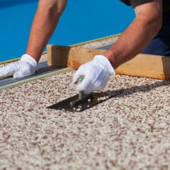 A person wearing white gloves is using a trowel to smooth the surface of a roof covered with small, white gravel-like material. A wooden plank and a blue sky are visible in the background.