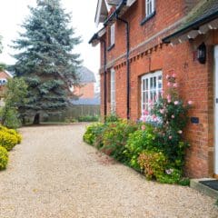 A charming brick house with white-framed windows and a white door. A gravel path leads to the entrance, flanked by vibrant flowers and shrubs. A tall evergreen tree stands in the yard, with neighboring houses visible in the background.