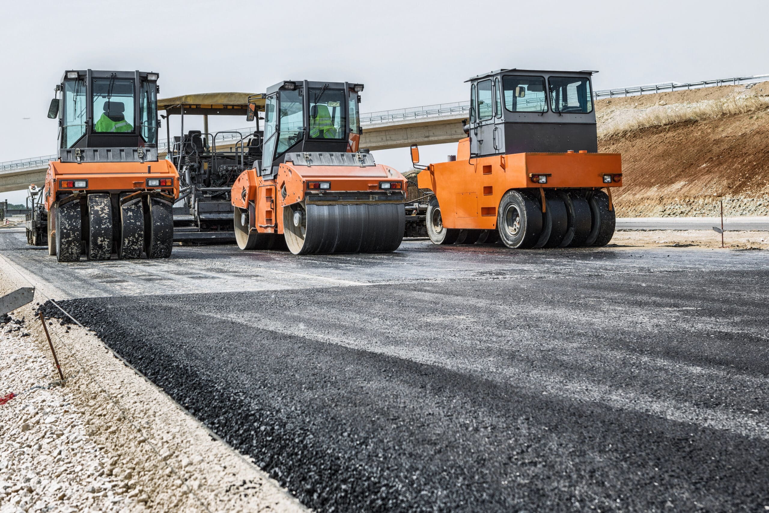 Three orange road rollers compact fresh asphalt on a highway under construction. The machinery is aligned side by side, ready to flatten the surface. An overpass is visible in the background, and the sky is clear.