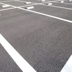 Empty parking lot with neatly painted white lines marking parking spaces. Asphalt surface is smooth and clean, with several wheel stops placed at the front of each space. Bright daylight casts subtle shadows across the area.