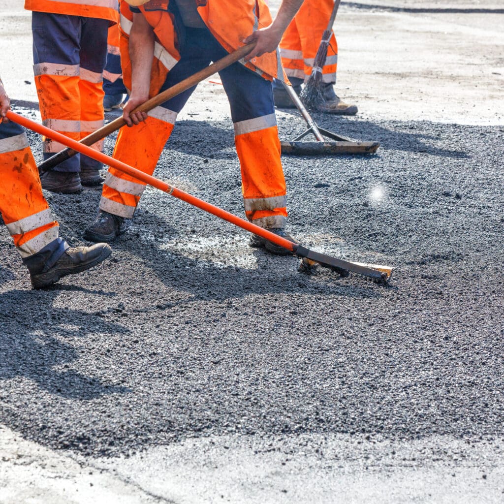 Construction workers in orange safety gear are using tools to spread asphalt on a road surface. The image shows a close-up of their legs and tools as they work on paving the road.