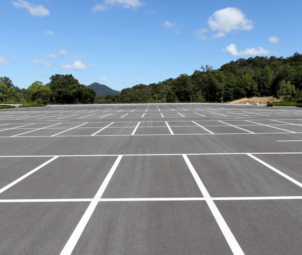 An empty, expansive parking lot with neatly painted white lines under a blue sky scattered with clouds. Trees and low hills are visible in the background.