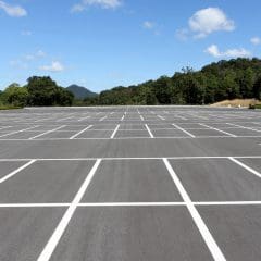 An empty, expansive parking lot with neatly painted white lines under a blue sky scattered with clouds. Trees and low hills are visible in the background.