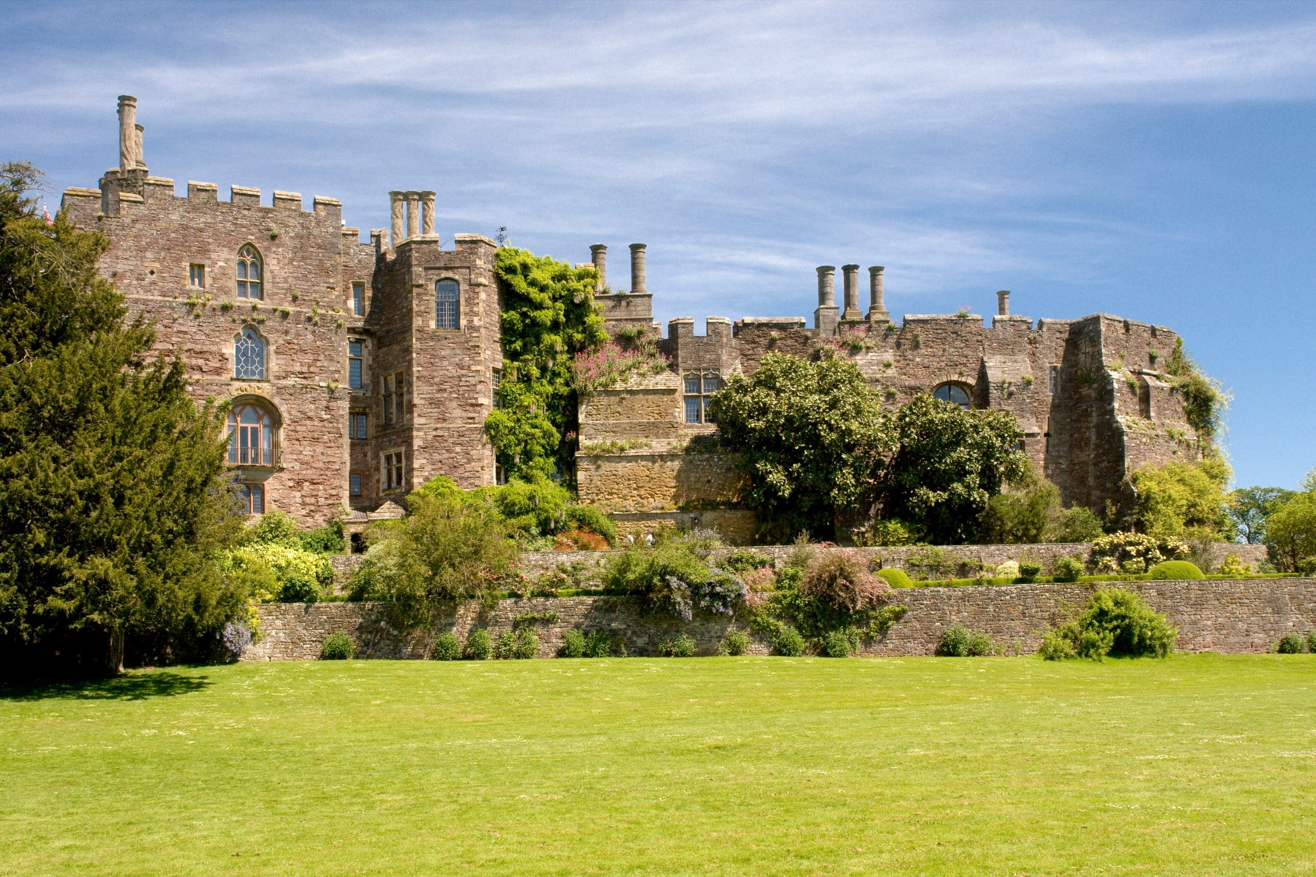 A historic stone castle with multiple turrets and chimneys is surrounded by lush greenery. It sits under a clear blue sky, with a well-kept lawn in the foreground.