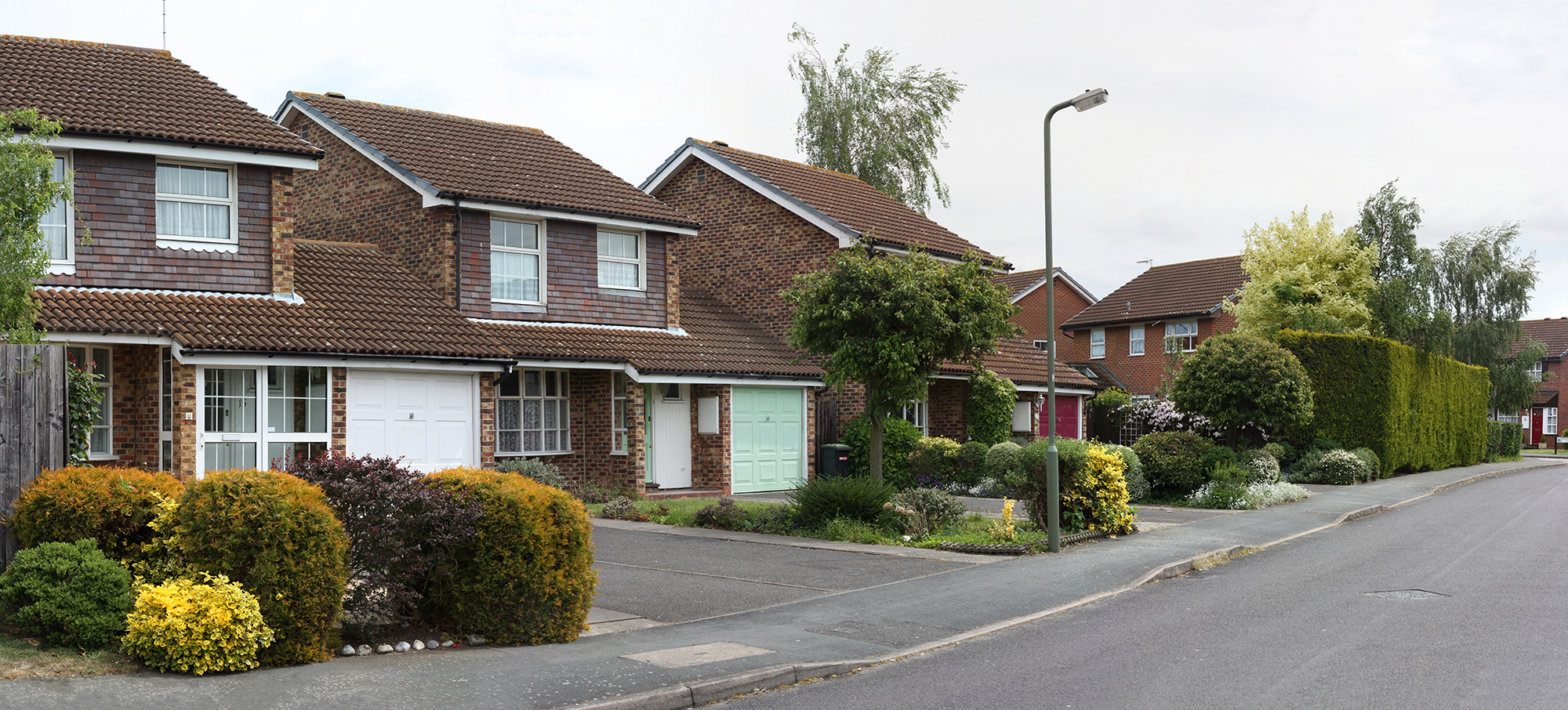 A suburban street with brick houses featuring brown tiled roofs. Each house has a garage and a small front garden with shrubs and trees. A streetlight stands on the sidewalk, and the street is quiet and empty.