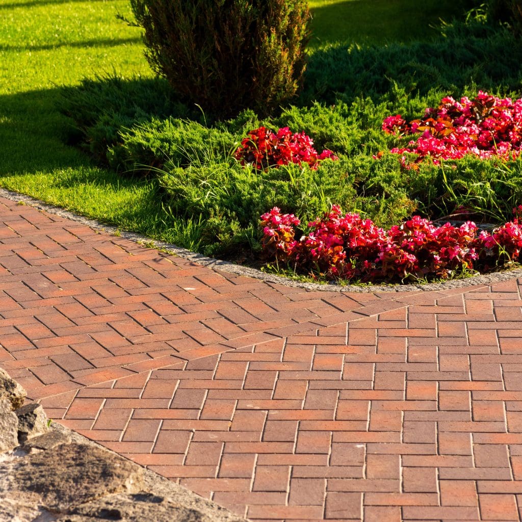 A brick pathway curves past a vibrant flowerbed filled with red blooms. The garden is lush with green foliage and neatly trimmed grass, bathed in warm sunlight.