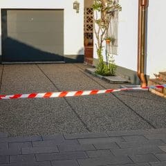 A driveway in front of a garage is blocked off with red and white caution tape. The ground has a speckled texture, and there are some plants near the entrance. The house exterior is white with a brown roof.