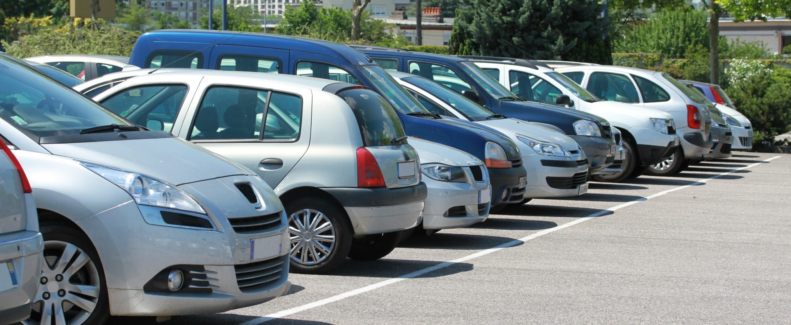 A row of various cars parked in an outdoor parking lot on a sunny day. Trees and buildings are visible in the background.