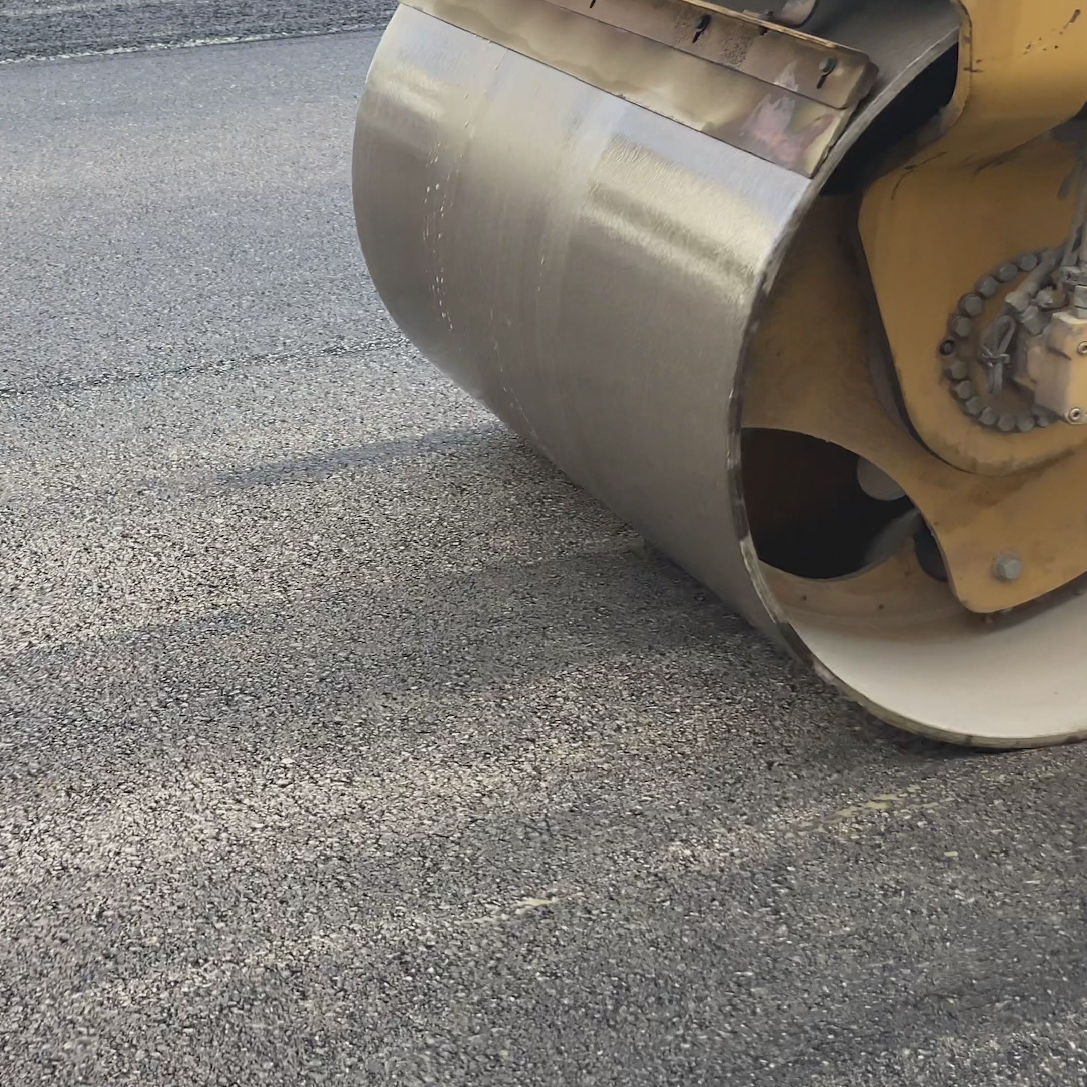 Close-up of a steamroller's metal drum rolling over newly laid asphalt on a road. The smooth surface indicates recent paving work. The roller is part of a large construction vehicle, visible partially in the image.