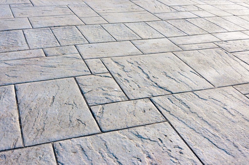 Close-up view of a textured stone pavement with a pattern of rectangular and square tiles. The surface has subtle natural variations in color, showcasing a mix of gray and light brown tones, creating a rustic and elegant appearance.