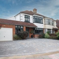 A two-story suburban house with a red brick and cream facade, featuring a front garden, paved driveway, and a garage. The sky is partly cloudy. Shrubs and trees border the property.