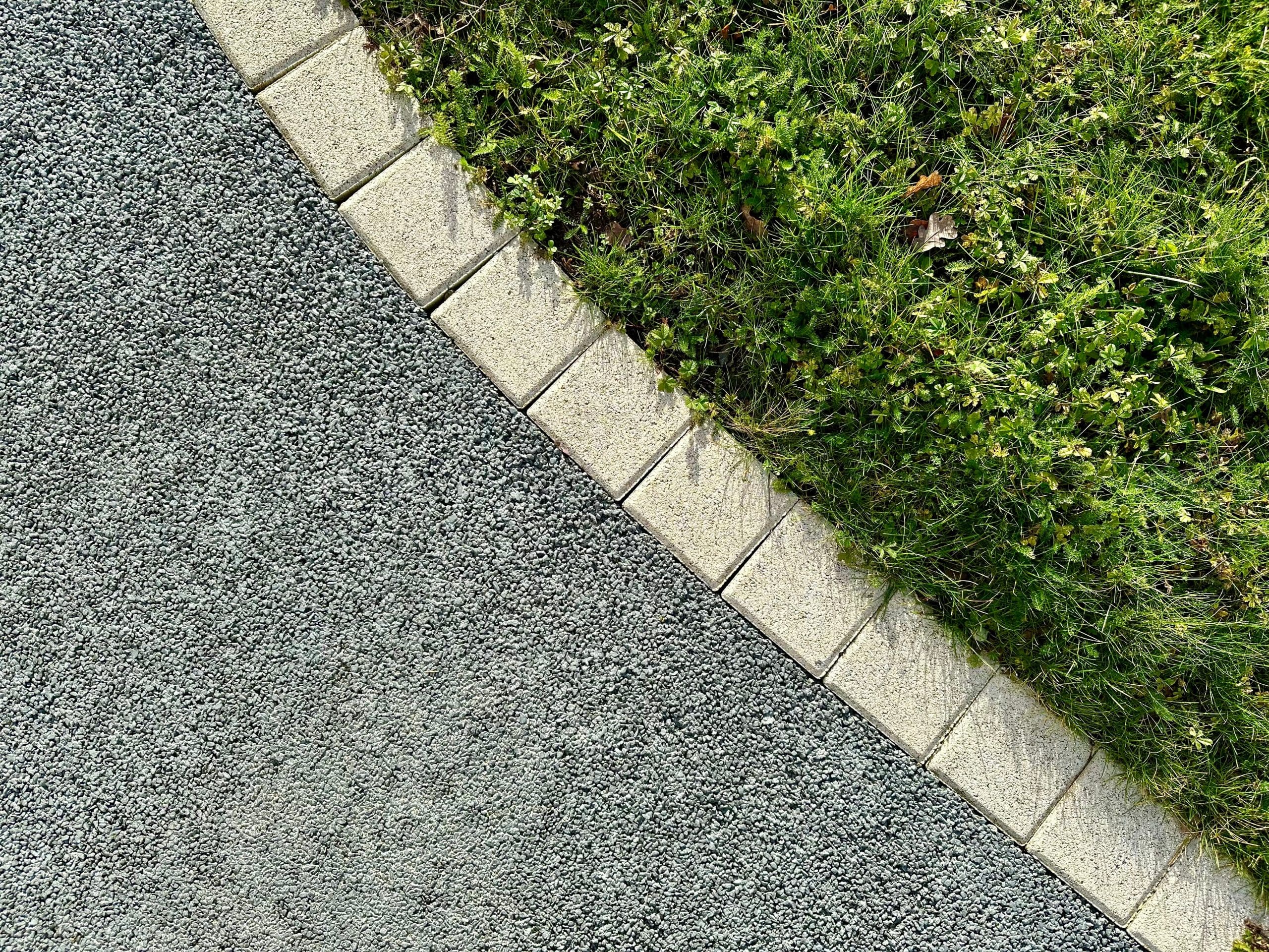 A top-down view of a curved border between a gray gravel path and a green grassy area. Beige stone tiles neatly separate the two zones, showing a clear contrast between the textured gravel and lush grass.