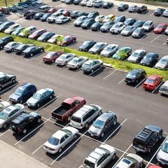 Aerial view of a large parking lot filled with numerous cars, organized into several rows. The lot is bordered by a few green, grass-covered areas and trees. The scene is well-lit, indicating daytime.