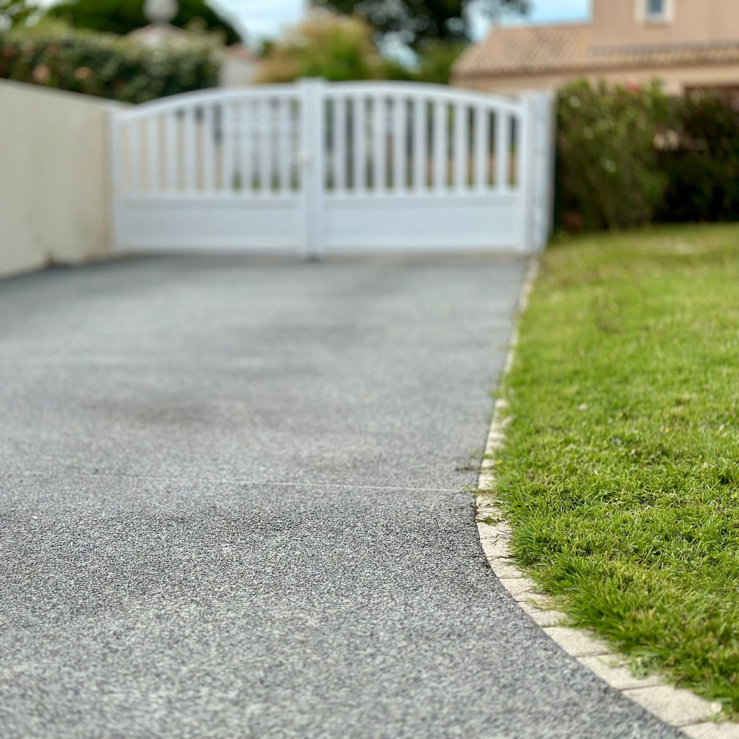 A curved, paved driveway leads to a closed white fence with vertical slats. Green grass and shrubbery border the driveway on one side, while a white wall lines the other. Neutral-colored houses are visible in the background under a cloudy sky.