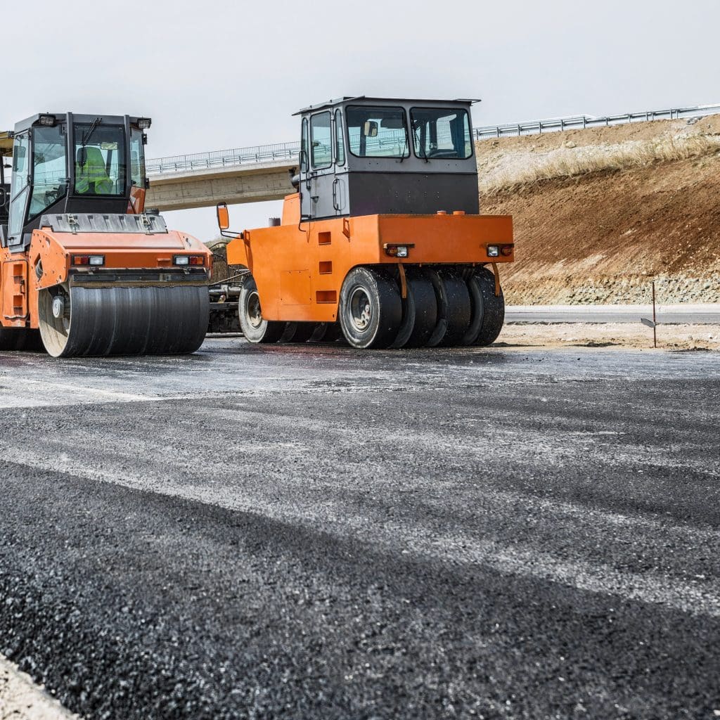 Two orange road rollers on a freshly paved asphalt road with a bridge in the background. The scene depicts construction work under a clear sky.