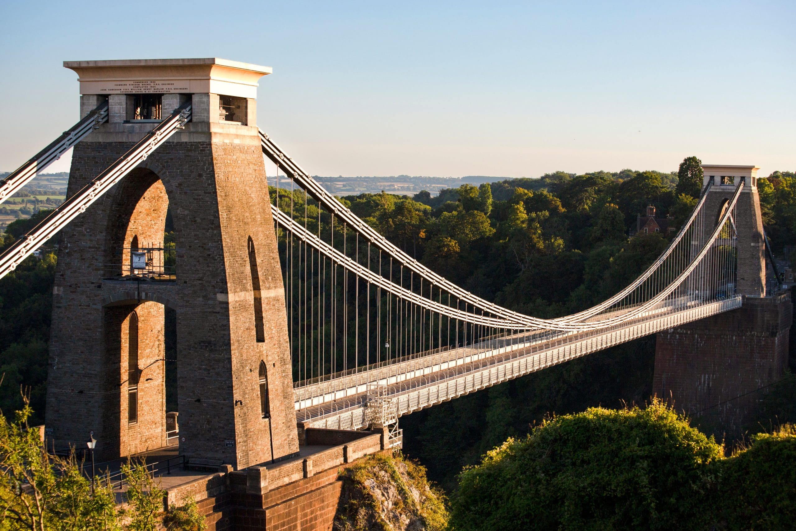 Suspension bridge with tall stone towers and long cables stretching across a river gorge, surrounded by lush green trees under a clear blue sky.