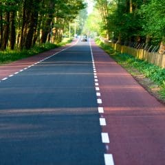 A scenic view of a straight, empty road bordered by dense green trees and grass on a bright day. The road has clear white lane markings and extends into the distance, where a single car is visible.