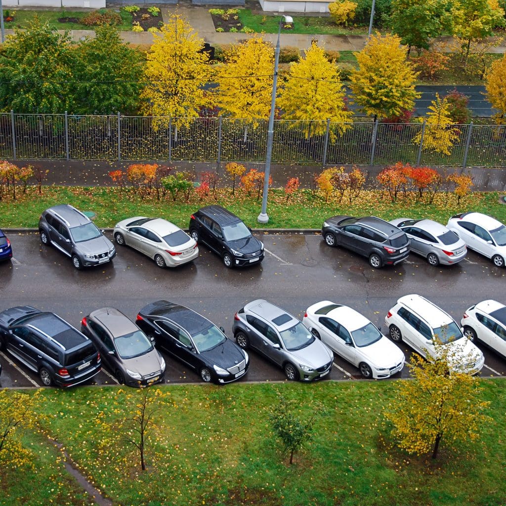 Aerial view of a parking lot with two rows of parked cars surrounded by trees with autumn leaves. The ground is wet, suggesting recent rain, and a fence separates the lot from a road lined with more colorful trees.