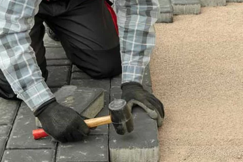 A person wearing gloves and a plaid shirt uses a rubber mallet to install interlocking paver bricks on a sandy base. The image shows hands working on aligning the dark gray pavers with rows already in place.