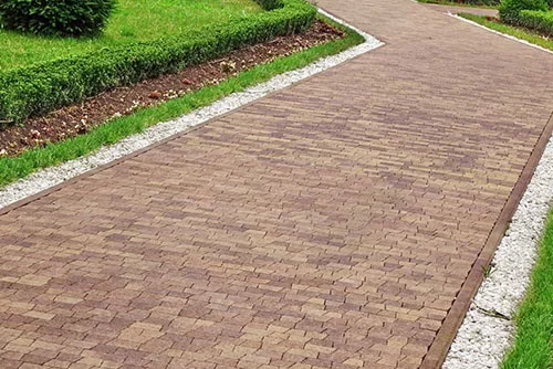 A narrow cobblestone pathway lined with small green bushes and grass on either side. The pathway features a border of white gravel, and the green vegetation appears well-maintained.