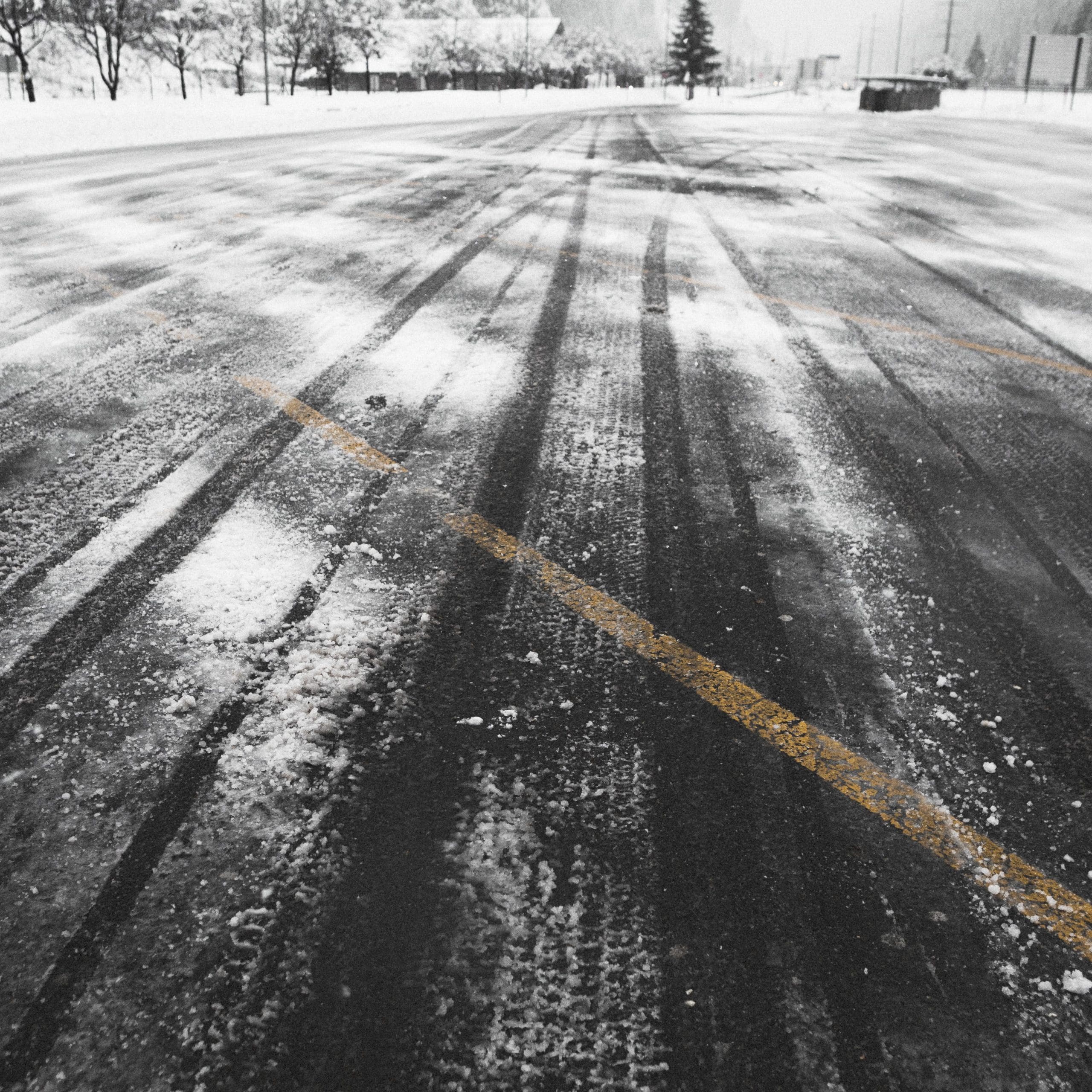 Snow-covered road with visible tire tracks and a faint yellow line. Snow-laden trees and a snowy landscape are in the background, creating a wintery scene.