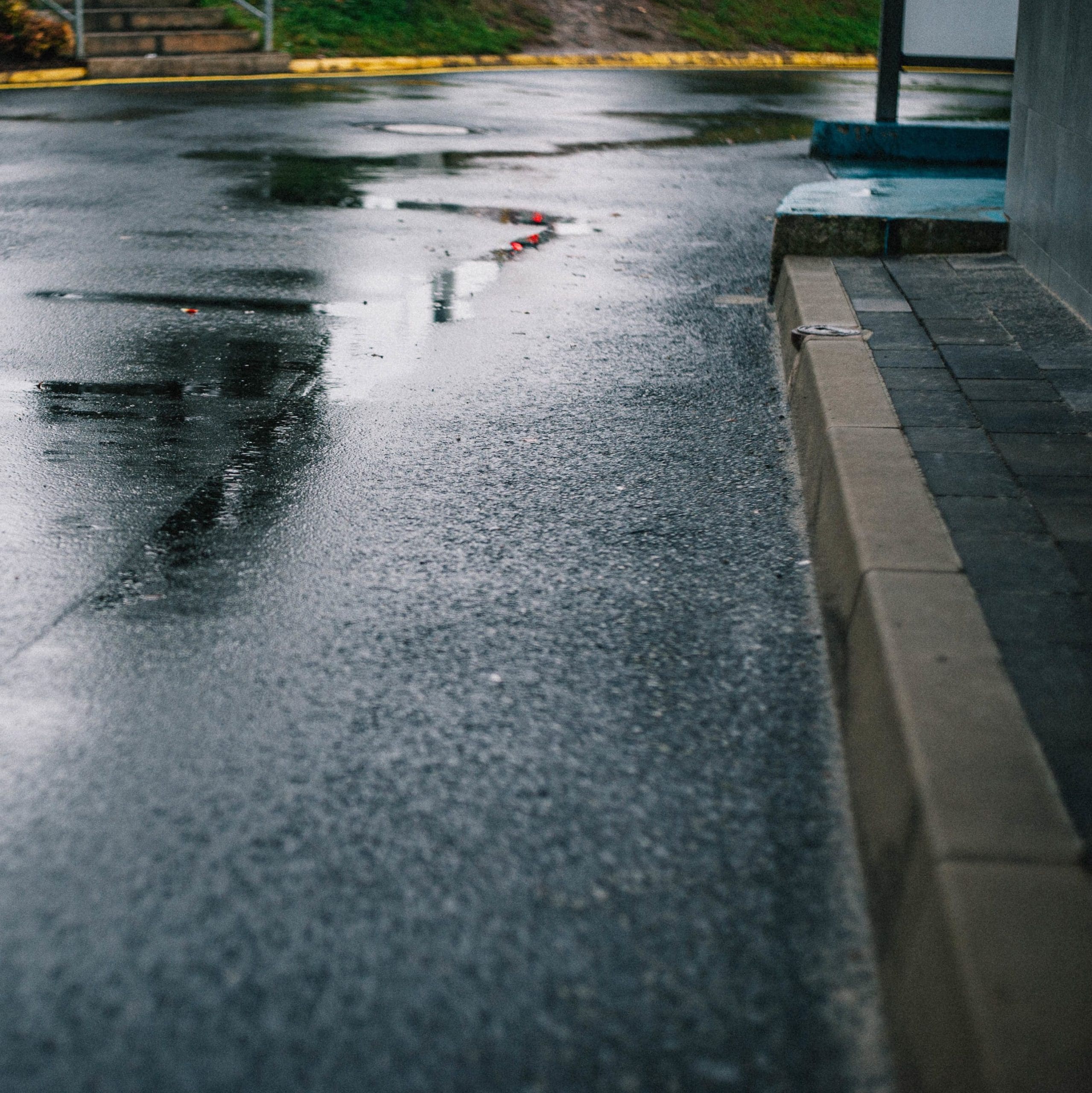 Wet pavement with light reflections on a rainy day. Curb and sidewalk visible on the right, with small puddles scattering across the road. Hints of greenery and a staircase are visible in the background.