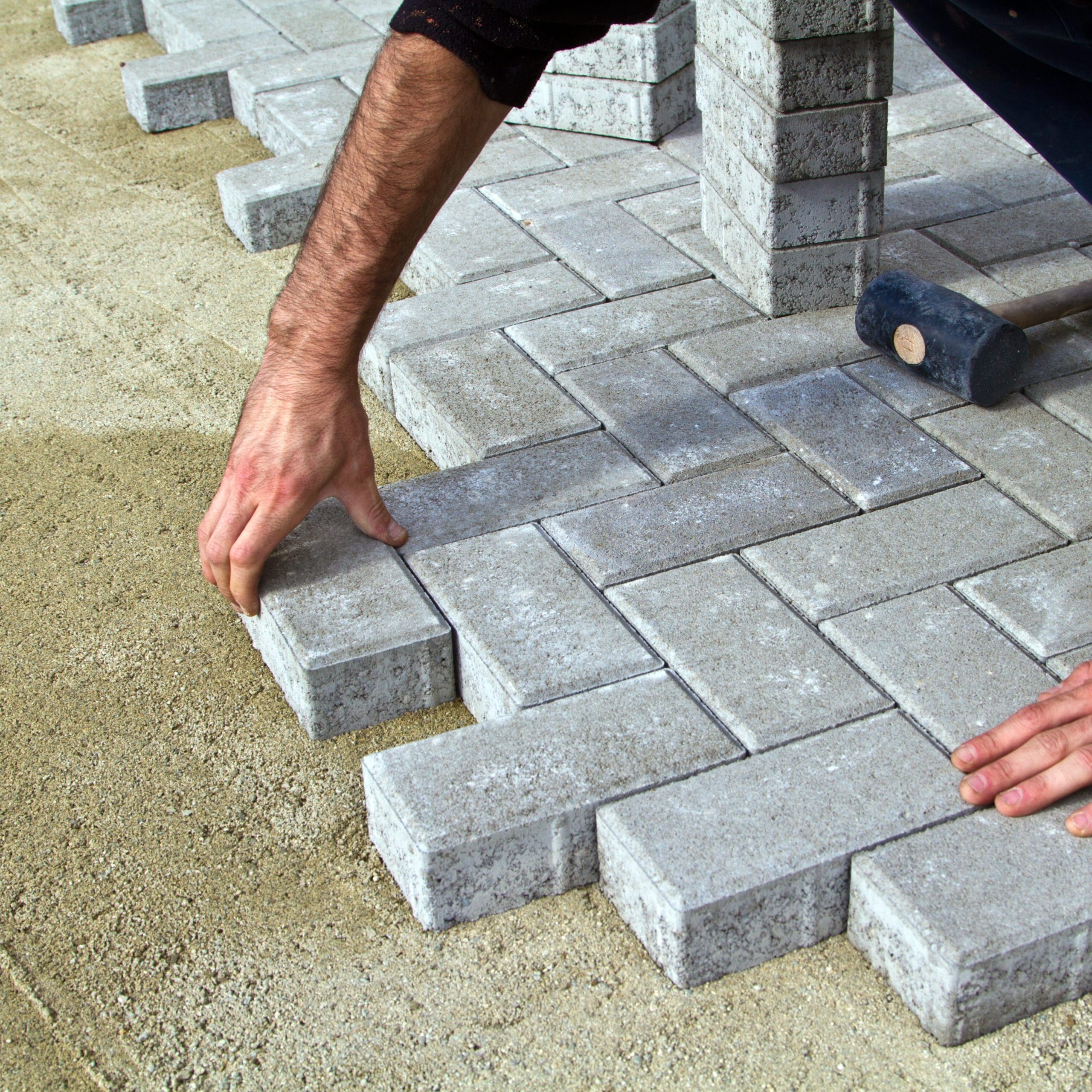 A person is laying gray rectangular paving stones in a pattern on a sandy surface. A rubber mallet rests nearby. The stones are tightly fitted together, forming a neat interlocking design.