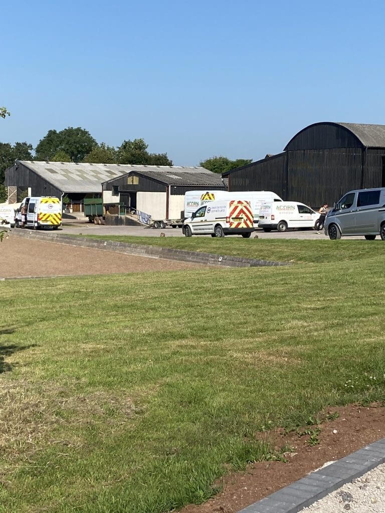 A rural scene with several work vans parked near farm buildings. The area is surrounded by greenery and the sky is clear.