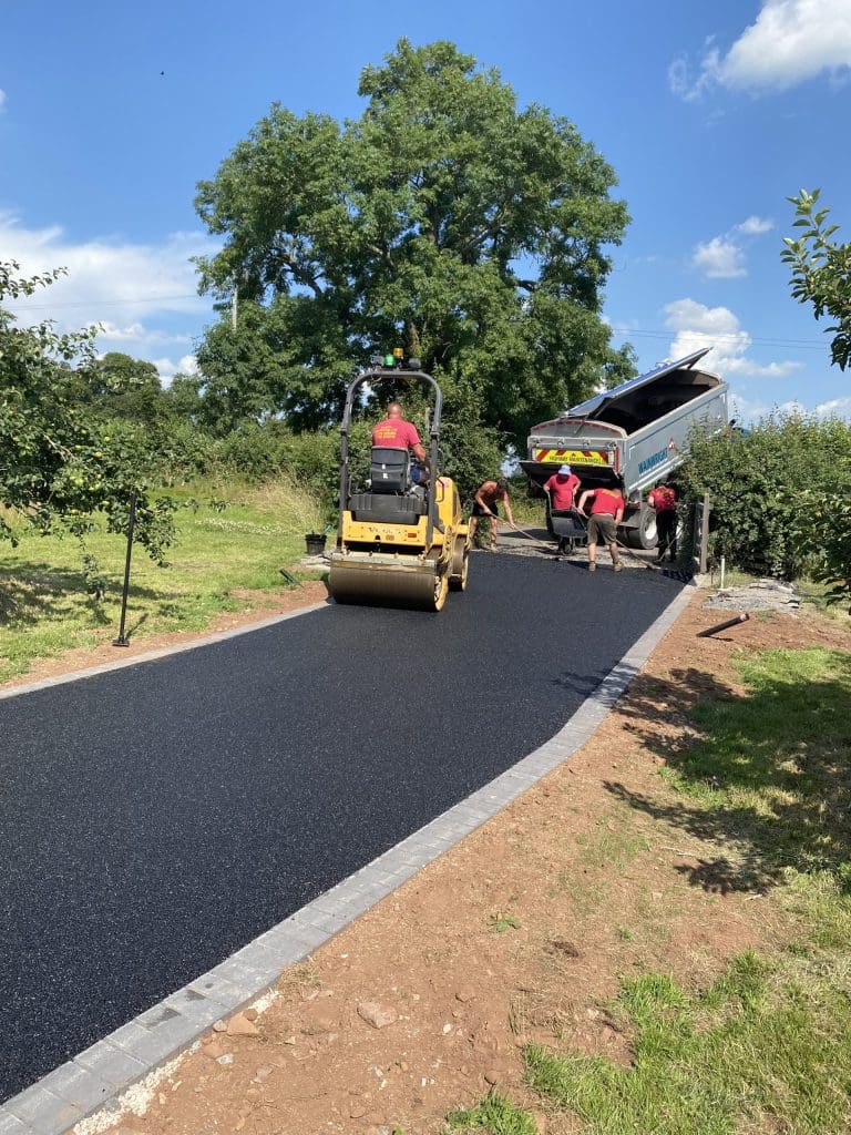 Workers are paving a driveway with fresh asphalt on a sunny day. A steamroller compacts the surface while a truck unloads more asphalt. Greenery surrounds the area, and the sky is clear blue with scattered clouds.