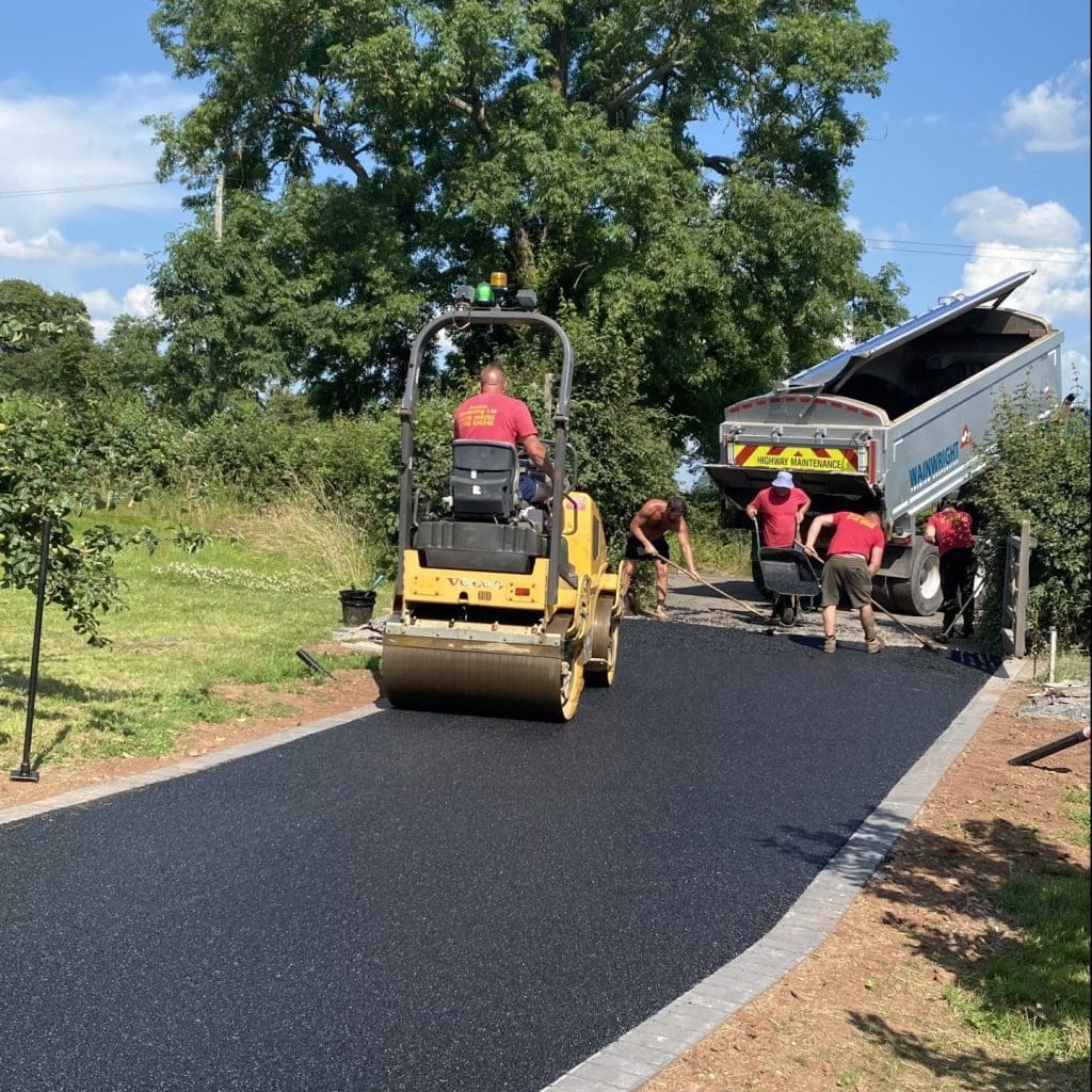 Workers are paving a driveway. A man operates a steamroller to flatten the asphalt while others spread it from a truck. The setting is outdoors with trees and greenery around. The sky is blue with a few clouds.