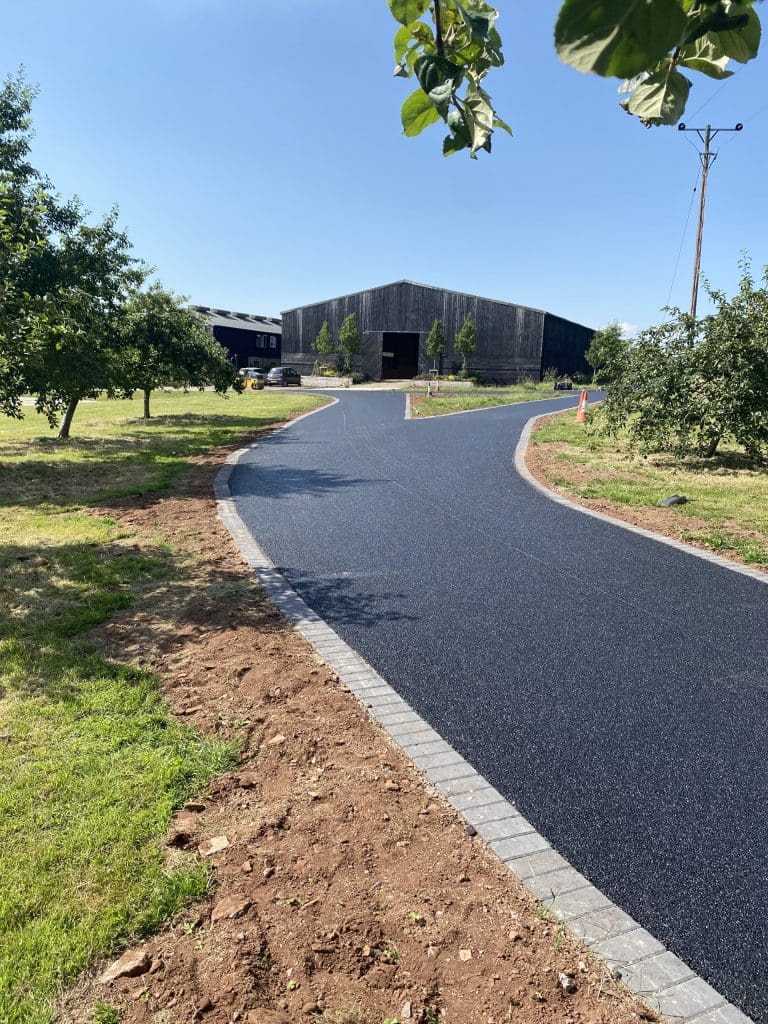 A newly paved asphalt path curves through a green area with trees, leading to a large wooden building in the distance. It's a sunny day, and the path is bordered by grass and some soil.