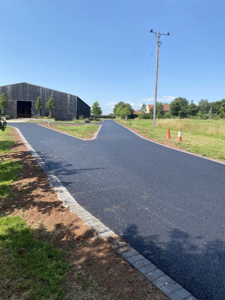 A freshly paved road splits into two paths in a rural area. Orange traffic cones are placed near the intersection. A wooden barn is visible on the left side, with green trees and grass surrounding the scene under a clear blue sky.