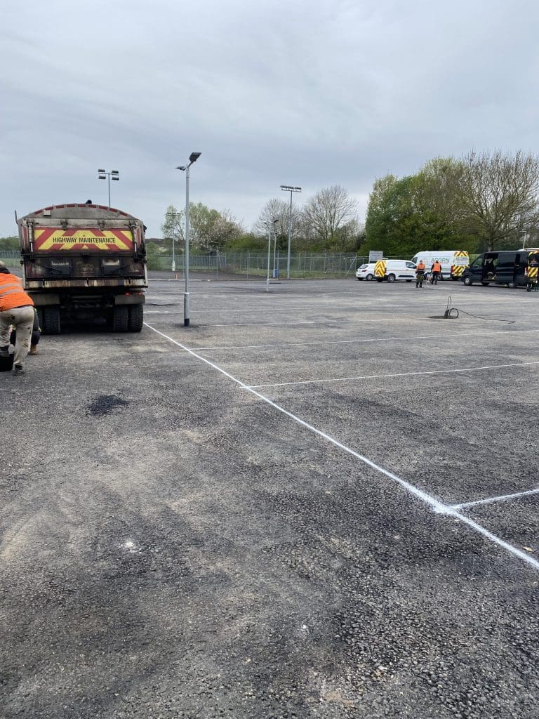 A parking lot under development, featuring several workers in orange vests, a truck with "Highway Maintenance" signage, and two white vans parked nearby. The pavement is marked with white lines, and trees are visible in the background.