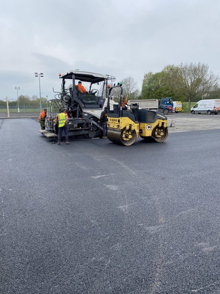 Construction workers operate an asphalt paver and roller on a freshly laid section of road. The area is surrounded by trees and utility vehicles are parked nearby. The sky is overcast.