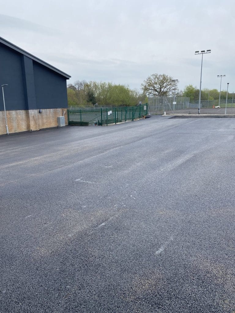 A newly paved, empty parking lot is shown with a building on the left. There are green fences and a few light poles in the background. Trees and overcast skies can be seen in the distance.