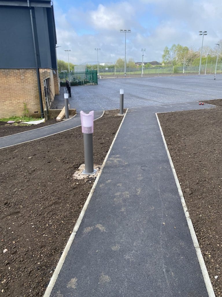 A newly paved walkway surrounded by dirt and installed lights, leading towards a gate. A building wall is on the left, with a cloudy sky and trees in the distance.