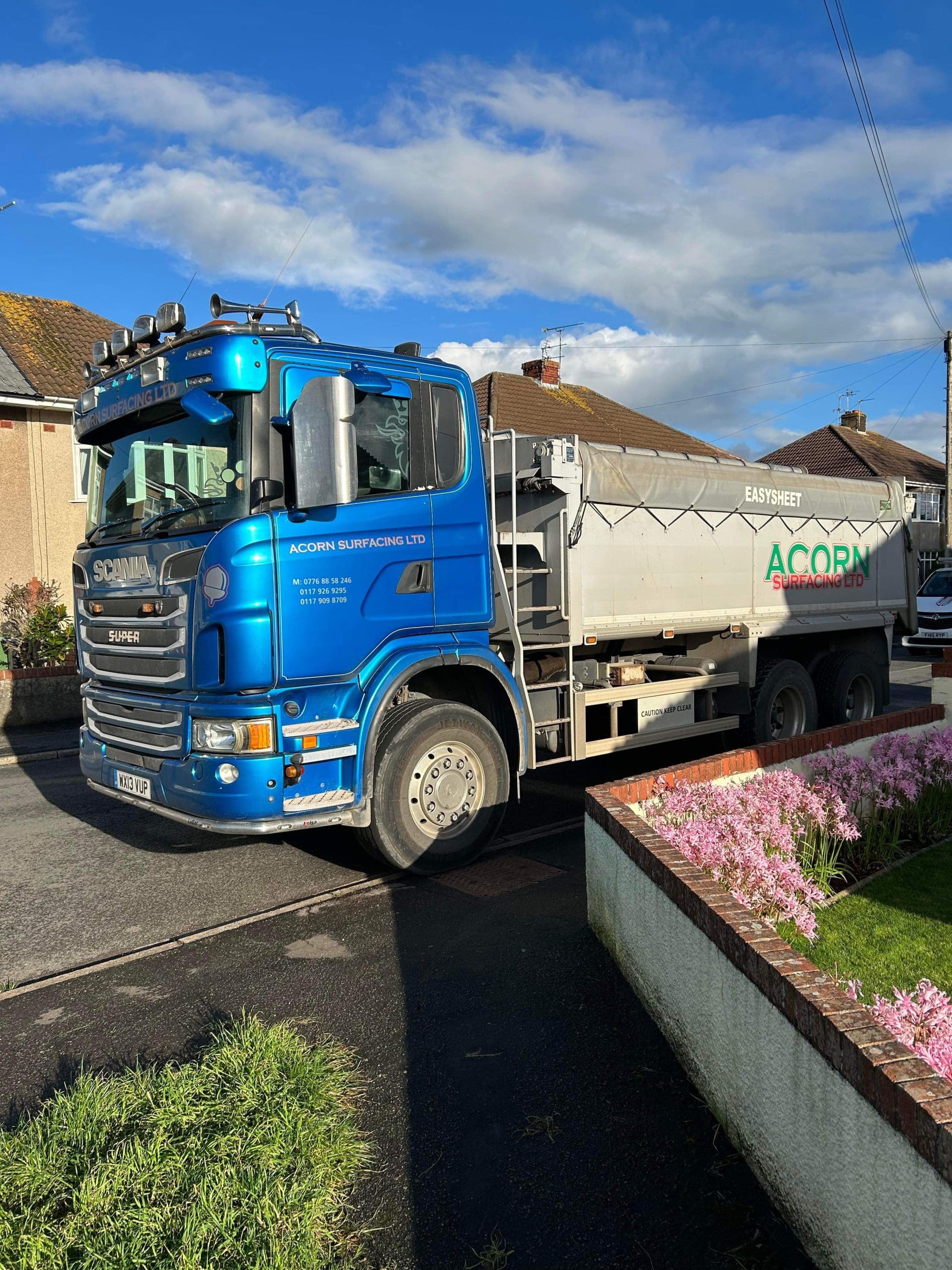 A blue Scania truck from Acorn Surfacing Ltd parked on a residential street. The truck has a silver EasySheet cover. Pink flowers and houses are visible under a partly cloudy sky.