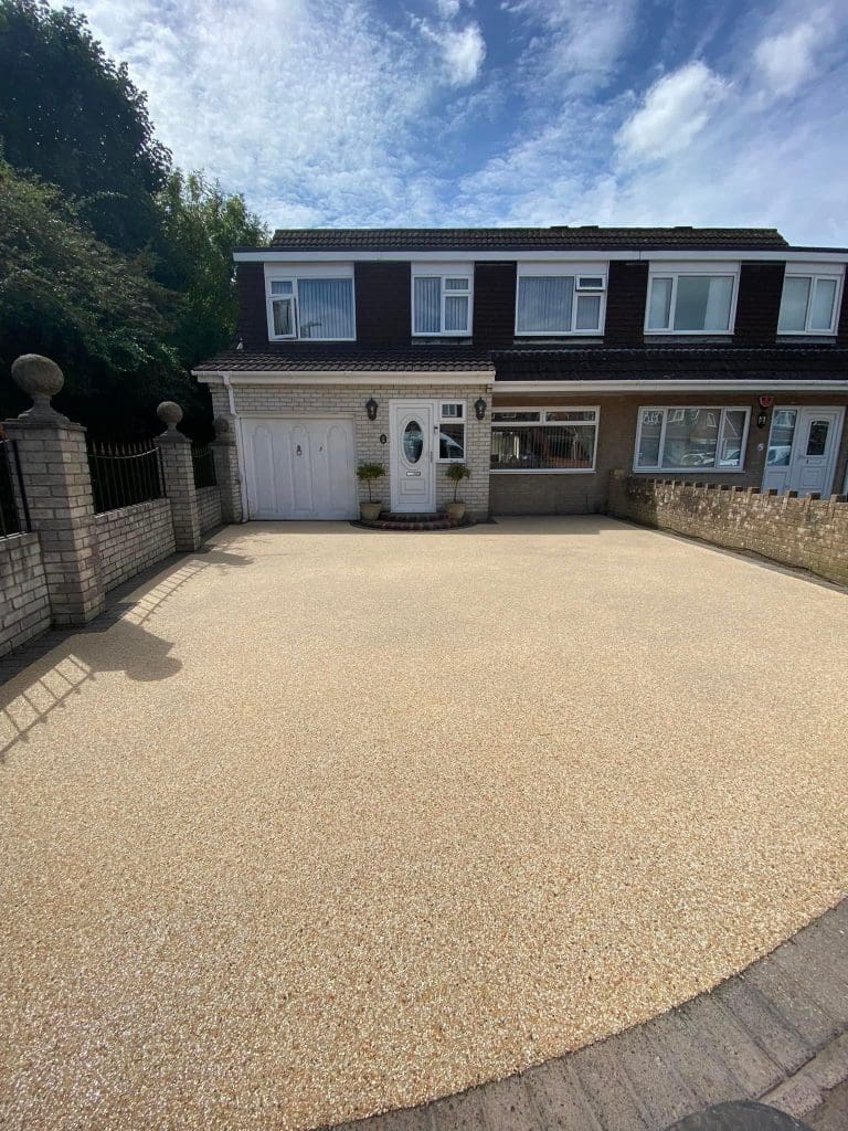 A semi-detached house with a newly surfaced driveway in beige gravel. The sky is partly cloudy, and there are trees and brick fences on the sides. A white garage door and front door with potted plants are visible.