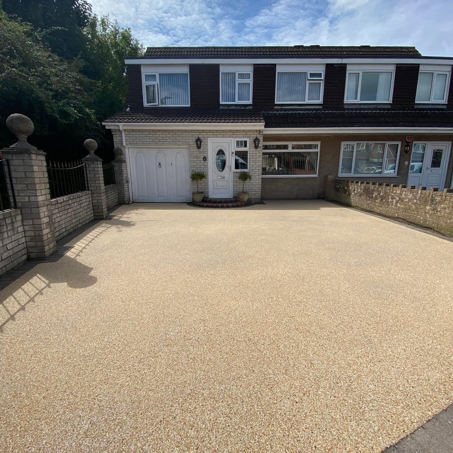 A semi-detached house with a white exterior, featuring a garage on the left and a white front door in the center. The driveway is freshly paved with light-colored gravel. A low brick wall with pillar accents borders the property.