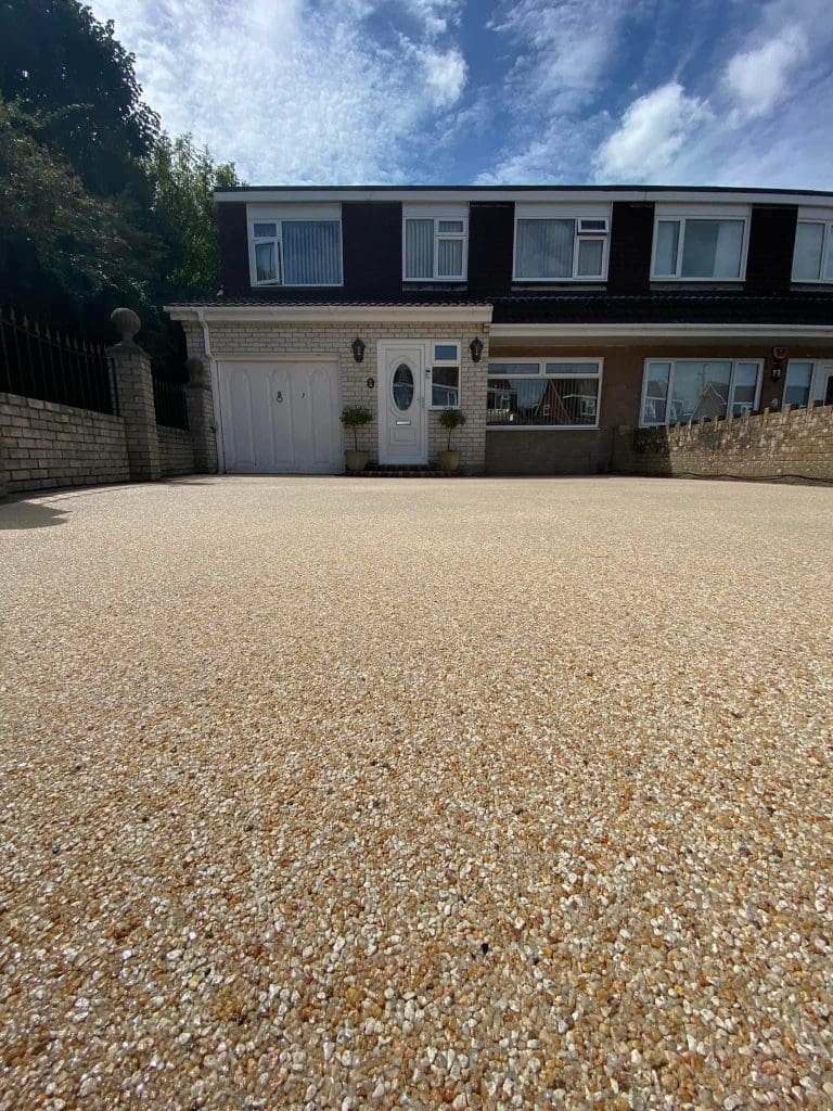 A suburban house with a large, smooth resin-bound driveway leading to a white garage and entrance door. The sky is partly cloudy, and greenery is visible to the left. The driveway has a textured, speckled appearance in shades of beige and brown.