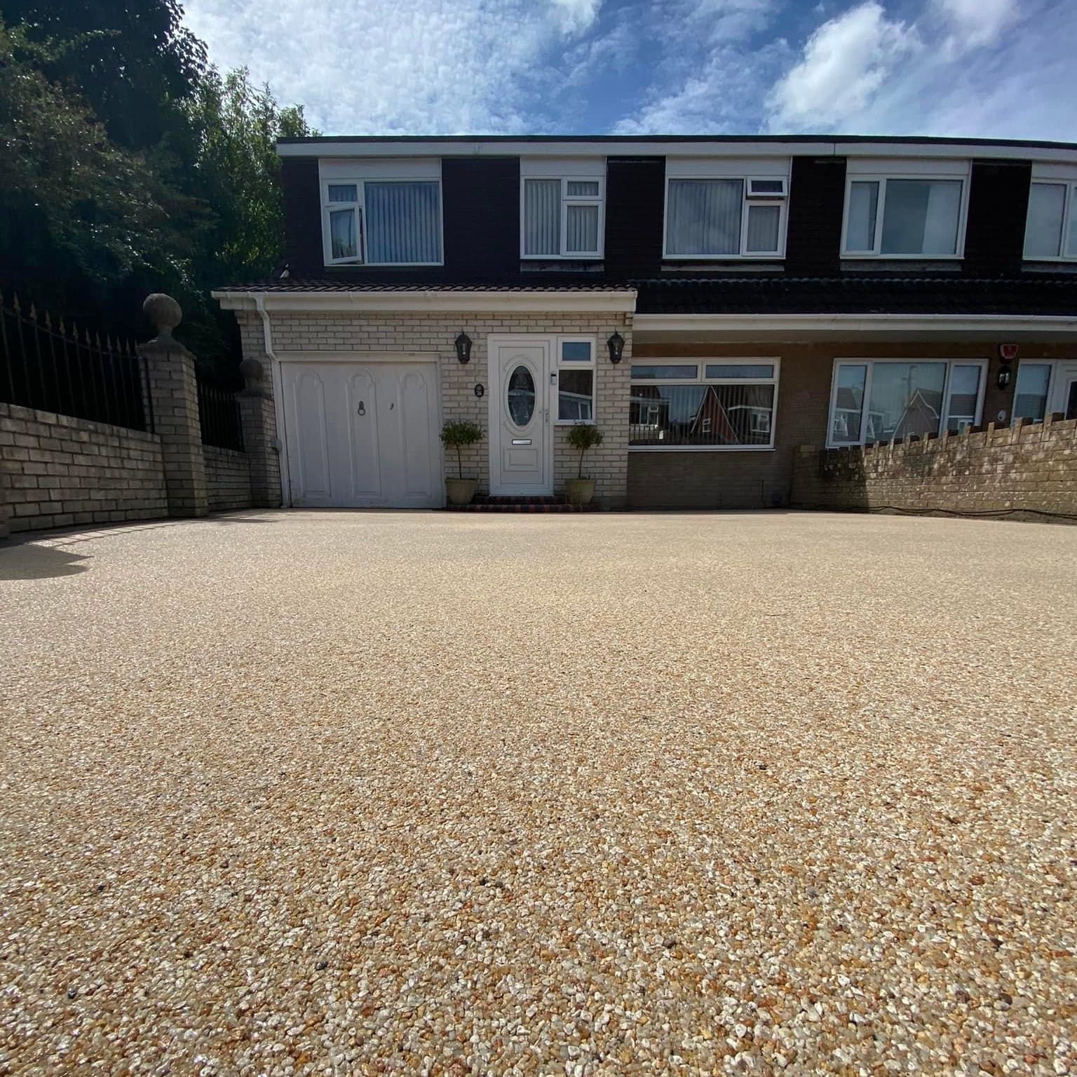 A two-story suburban house with white and brown exterior. A large, smooth pebble-finished driveway leads up to the garage. The sky is partly cloudy, and there are some trees on the left side.