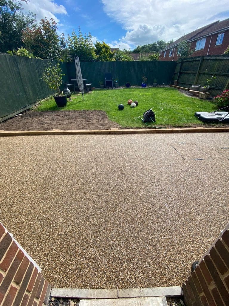 View of a backyard with a new gravel patio area in the foreground. The grass lawn has scattered sports balls and a few potted plants. The yard is enclosed by a wooden fence, with houses visible in the background under a partly cloudy sky.