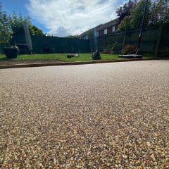 A close-up of a freshly laid resin driveway with a textured, pebbled surface. In the background, there's a grassy garden area with a basketball hoop, potted plants, and a wooden fence under a partly cloudy sky.