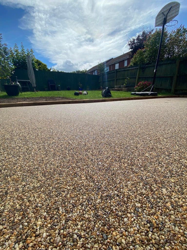 A close-up of a freshly laid resin driveway with a textured, pebbled surface. In the background, there's a grassy garden area with a basketball hoop, potted plants, and a wooden fence under a partly cloudy sky.