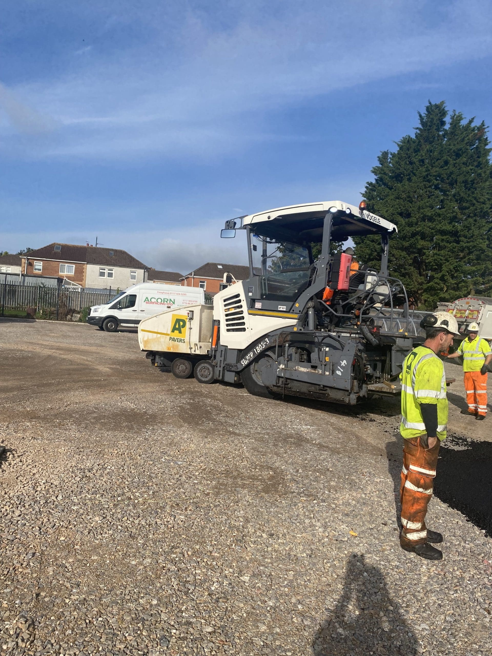 Construction workers in safety gear operate a large machine laying asphalt on a road. Nearby, there's a parked vehicle with the word "ACORN" visible. Houses and trees are in the background under a clear sky.