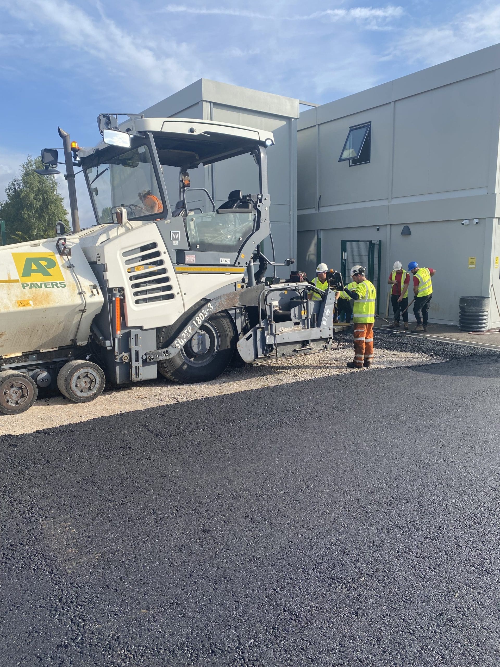 Workers in high-visibility clothing operate a paving machine, laying new asphalt on a road. Several other workers stand near a building in the background, observing the process. The scene is under a clear blue sky.