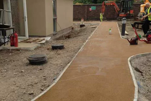 Construction site with a newly paved path leading to a building entrance. Construction equipment and workers are visible in the background. There are materials like pipes and a fire extinguisher along the sides of the path.