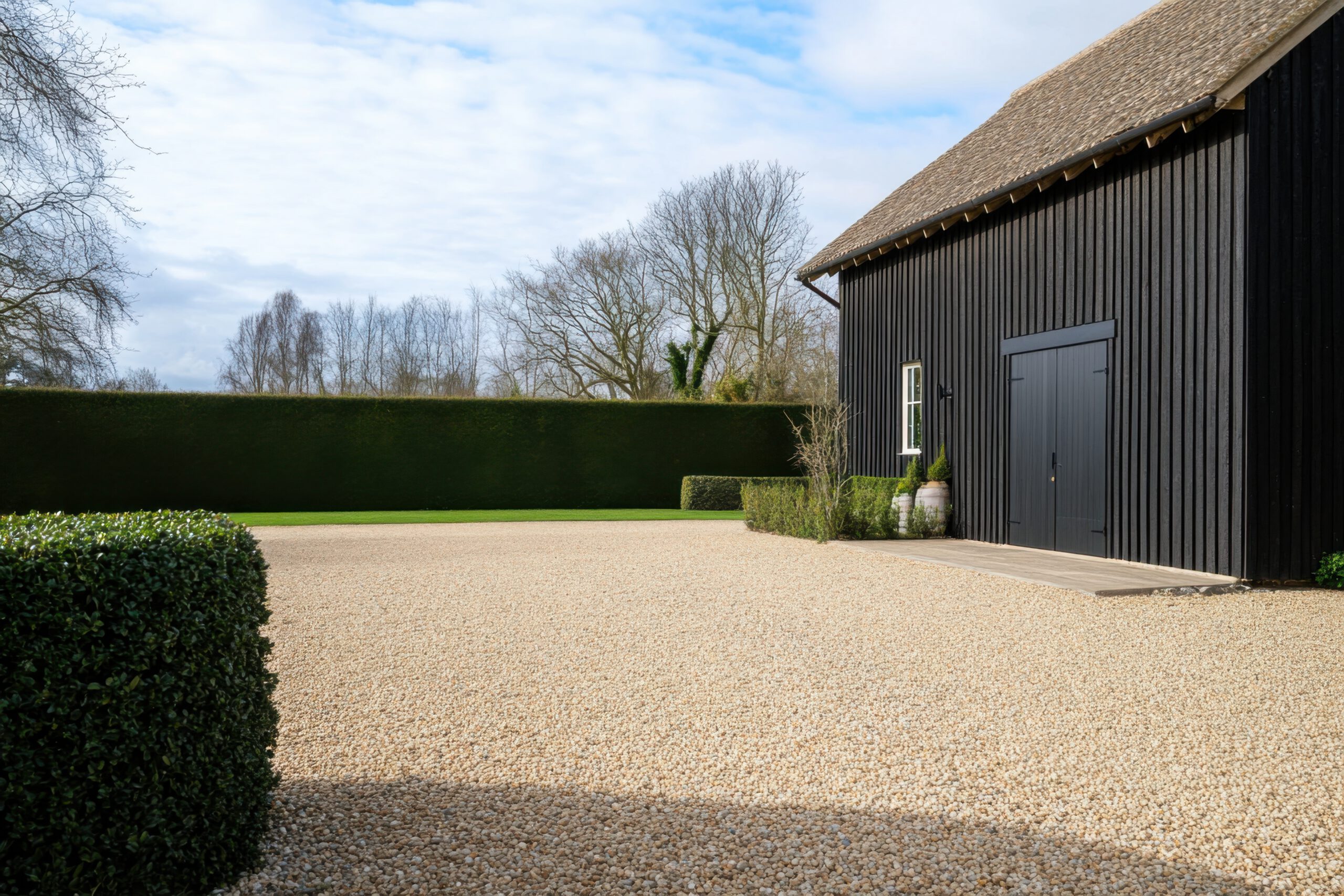 A modern black barn with double doors sits beside a gravel driveway, bordered by neatly trimmed hedges and lush green lawn, with bare trees and a cloudy sky in the background.