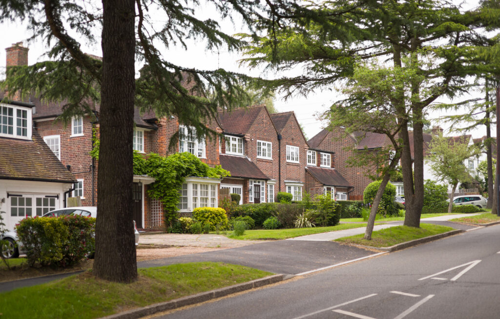 A quiet suburban street lined with brick houses, neatly trimmed hedges, and tall green trees. Sidewalks run along the houses, and a few parked cars are visible in driveways and on the street.