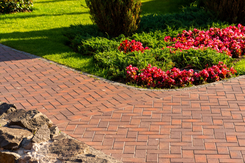 Red brick pathway with a corner, bordered by green grass, colorful red and pink flowers, and small shrubs in a landscaped garden. Rocks are visible along the edge of the walkway.